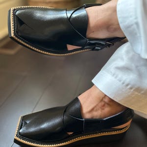 May include: Black leather Peshawari sandals with a square toe and a tan-coloured sole. The sandals have a strap design across the top. The shoes are a traditional style and are worn by someone wearing white trousers.