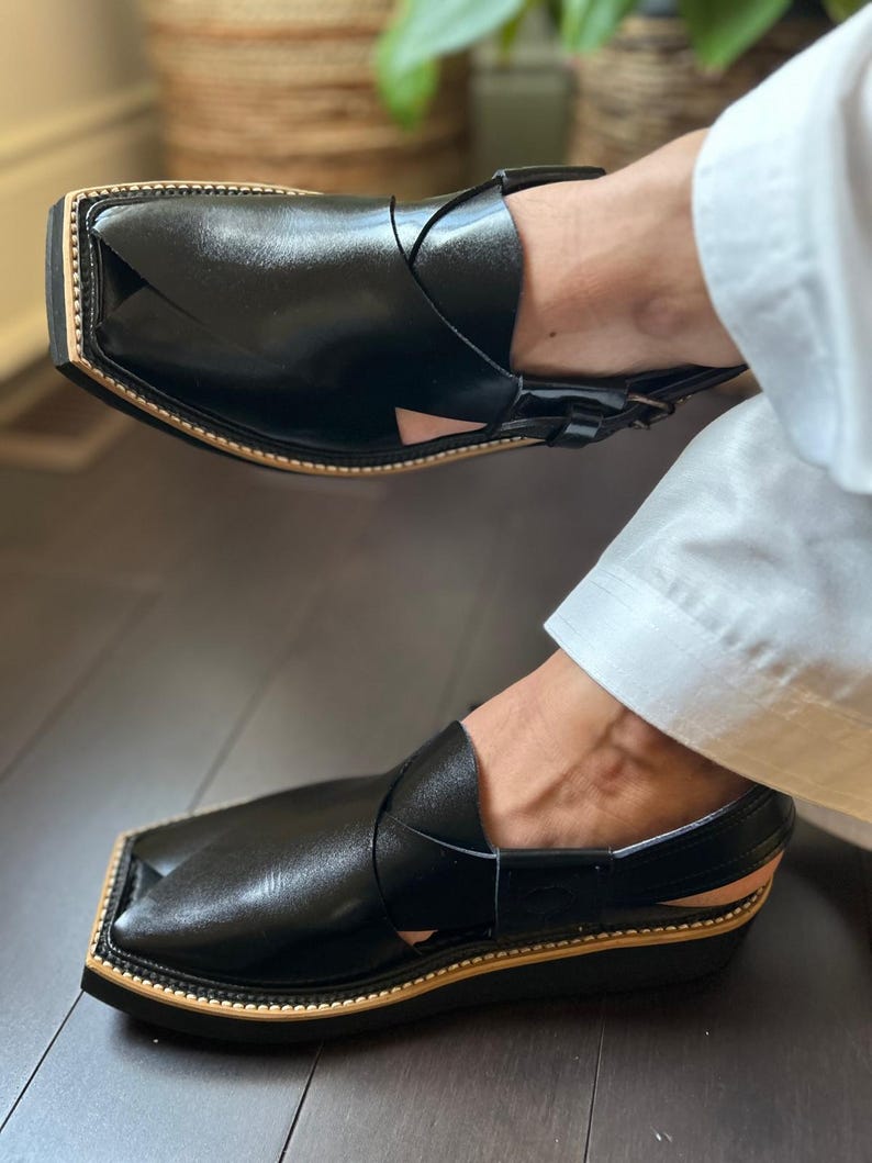 May include: Black leather Peshawari sandals with a square toe and a tan-coloured sole. The sandals have a strap design across the top. The shoes are a traditional style and are worn by someone wearing white trousers.