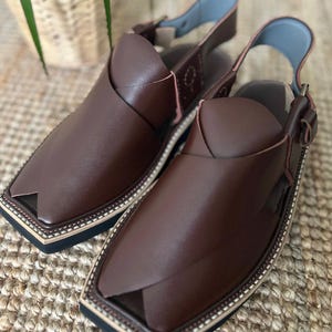 May include: A pair of brown leather Peshawari sandals. These open-toe sandals feature a square toe design, an ankle strap, and a decorative stitched border. The sandals are displayed on a woven surface.