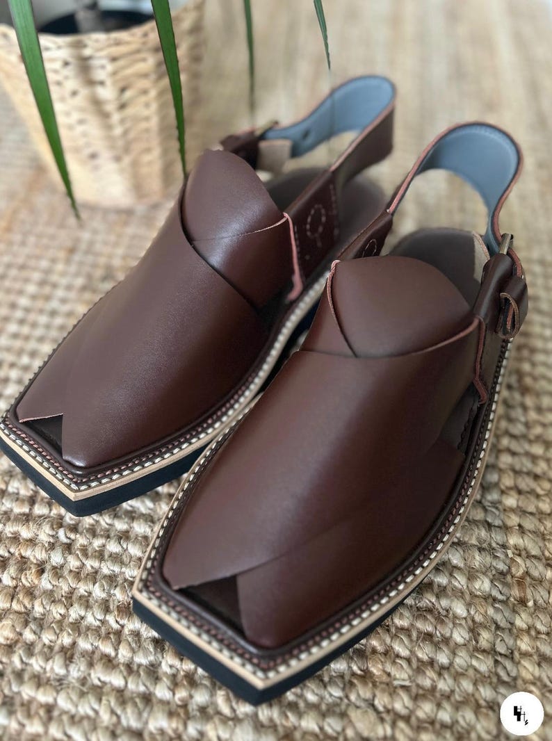 May include: A pair of brown leather Peshawari sandals. These open-toe sandals feature a square toe design, an ankle strap, and a decorative stitched border. The sandals are displayed on a woven surface.