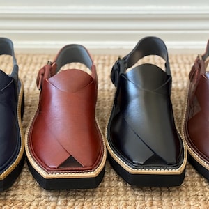 May include: Four pairs of leather Peshawari sandals in navy blue, burgundy, black, and brown. Each sandal features a strap, a buckle, and a thick sole with a light-coloured trim.