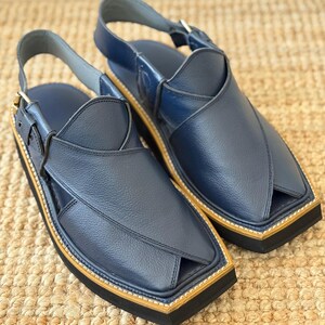 May include: A pair of navy blue leather sandals. The sandals have a unique design with a strap around the ankle and a buckle. The soles are black with a tan border. The sandals are presented on a woven surface.