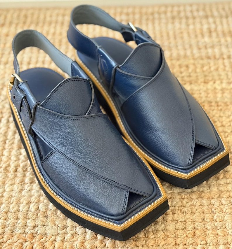 May include: A pair of navy blue leather sandals. The sandals have a unique design with a strap around the ankle and a buckle. The soles are black with a tan border. The sandals are presented on a woven surface.