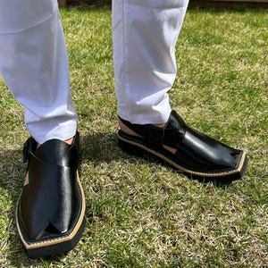 May include: Black leather Peshawari sandals with a square toe and a tan border. The sandals have a strap across the top and a buckle closure. The shoes are worn with white trousers on a grassy surface.