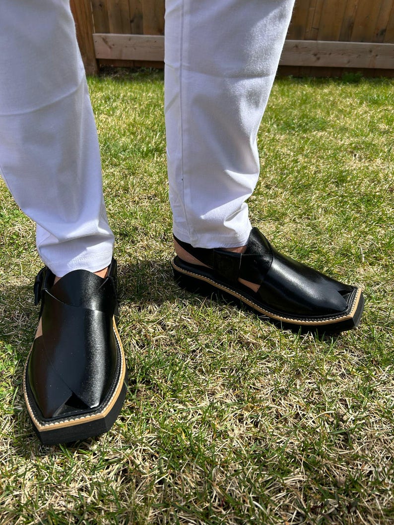 May include: Black leather Peshawari sandals with a square toe and a tan border. The sandals have a strap across the top and a buckle closure. The shoes are worn with white trousers on a grassy surface.