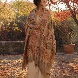 May include: A beige shawl featuring a multi-colored patchwork design with intricate embroidery in shades of red, gold, and brown. The rectangular wrap has a fringed hem and is draped over a plain beige top and wide-leg trousers in a matching tone.