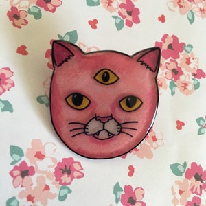 Pode incluir: Um alfinete em forma de gato rosa com três olhos amarelos e um contorno preto. O gato tem uma boca pequena com duas presas e bigodes. O alfinete está sobre um fundo floral com flores rosa e brancas.