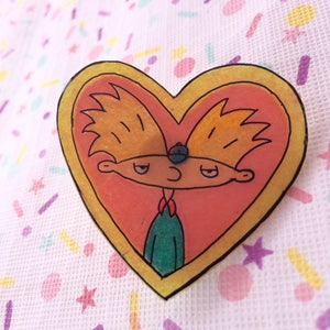 May include: A heart-shaped pin featuring a cartoon character with blonde hair and a blue hat. The character is inside a yellow border, with a peach-colored background. The pin is on a white fabric with colorful confetti.