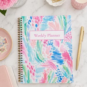 May include: A spiral-bound weekly planner featuring a colorful abstract watercolor pattern in shades of pink, blue, and green. The cover displays the text Weekly Planner centered within a white rectangular box with a thin pink border.