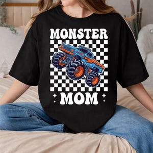 Puede incluir: Camiseta negra con un dise&ntilde;o gr&aacute;fico de un cami&oacute;n monstruo azul y naranja sobre un fondo a cuadros. El texto "MONSTER MOM" se muestra arriba y abajo del cami&oacute;n en letras blancas estilizadas.