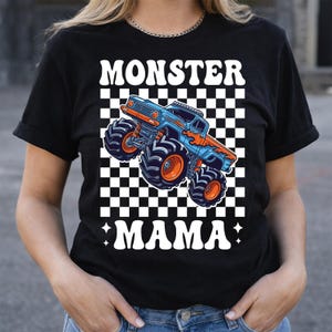 Op de afbeelding: Zwart T-shirt met een blauw en oranje monster truck ontwerp op een geruite achtergrond. De tekst "MONSTER MAMA" staat in witte, gestileerde letters. Het ontwerp bevat vlammen en grote banden.