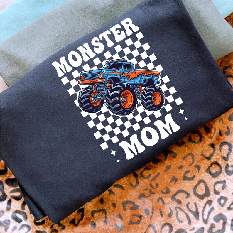 Puede incluir: Una camiseta negra con las palabras "MONSTER MOM" en blanco, junto con un gr&aacute;fico de un cami&oacute;n monstruo azul y naranja sobre un fondo a cuadros. La camiseta est&aacute; doblada y superpuesta con otras camisetas.