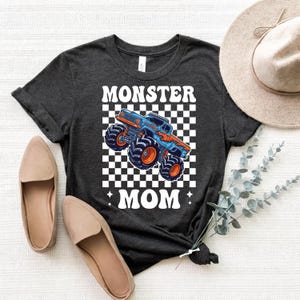 Puede incluir: Camiseta gris oscuro con un gr&aacute;fico de un monster truck azul y naranja sobre un fondo a cuadros, con las palabras "MONSTER MOM" en blanco. La camiseta se combina con bailarinas beige y un sombrero de ala ancha.