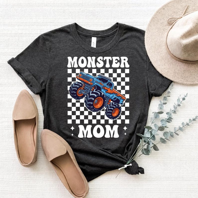 Puede incluir: Camiseta gris oscuro con un gr&aacute;fico de un monster truck azul y naranja sobre un fondo a cuadros, con las palabras "MONSTER MOM" en blanco. La camiseta se combina con bailarinas beige y un sombrero de ala ancha.