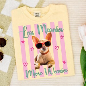 Puede incluir: Camiseta amarilla clara con un gráfico de un chihuahua con gafas de sol rojas. La camiseta tiene rayas rosas y amarillas con el texto "Less Meanies More Weenies" en escritura turquesa. También presenta corazones y estrellas rosas.
