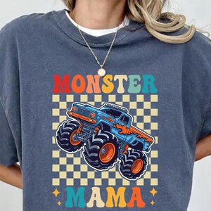 May include: A blue crew neck t-shirt featuring a colorful graphic of a monster truck. The design includes the words "MONSTER MAMA" in orange, teal, and yellow, set against a checkered background.