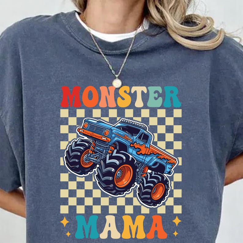 May include: A blue crew neck t-shirt featuring a colorful graphic of a monster truck. The design includes the words "MONSTER MAMA" in orange, teal, and yellow, set against a checkered background.