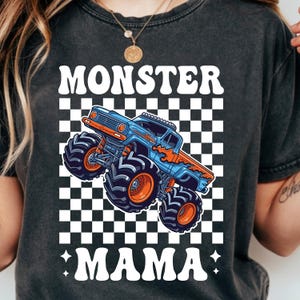 Op de afbeelding: Donkergrijs t-shirt met een blauw en oranje monster truck ontwerp op een zwart-wit geblokte achtergrond. De tekst "MONSTER MAMA" staat boven en onder de truck. De truck heeft grote banden.