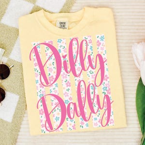 以下が含まれることがあります： ピンク色の「Dilly Dally」の文字が入った、薄い黄色のTシャツ。テキストはTシャツの中央に配置され、ピンク、青、白の花柄の縦縞模様があります。Tシャツはクルーネックで半袖です。