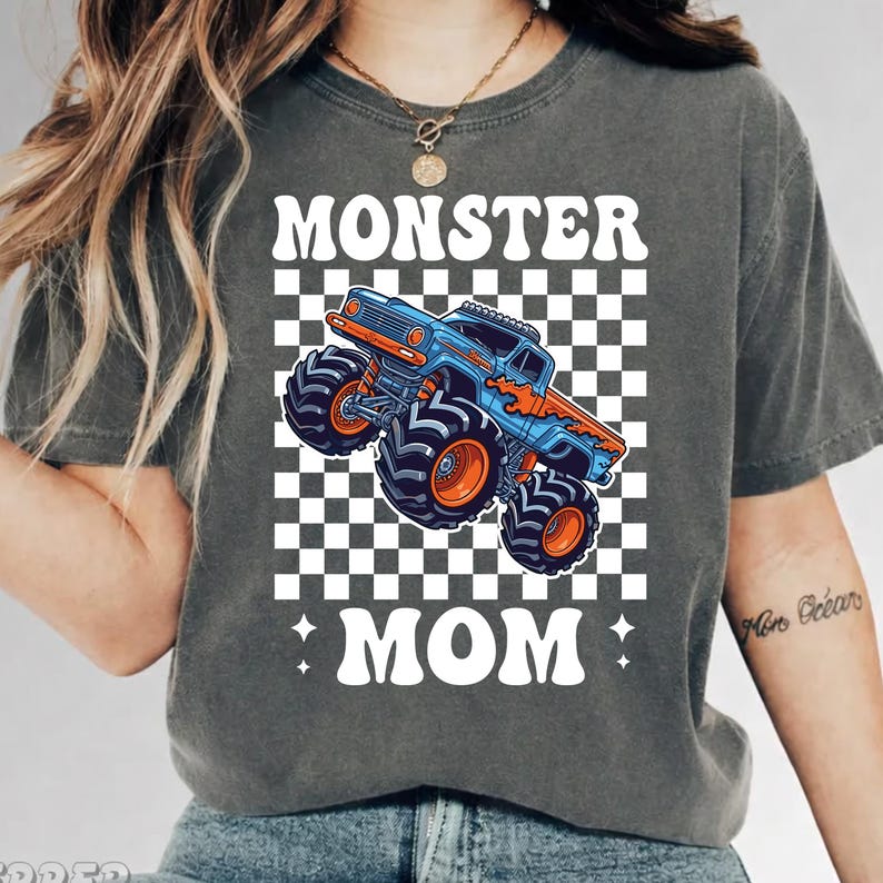 Puede incluir: Camiseta gris oscuro con un dise&ntilde;o de cami&oacute;n monstruo azul y naranja sobre un fondo a cuadros. El texto "MONSTER MOM" est&aacute; impreso encima y debajo del cami&oacute;n en letras blancas estilizadas. La camiseta tiene cuello redondo y mangas cortas.