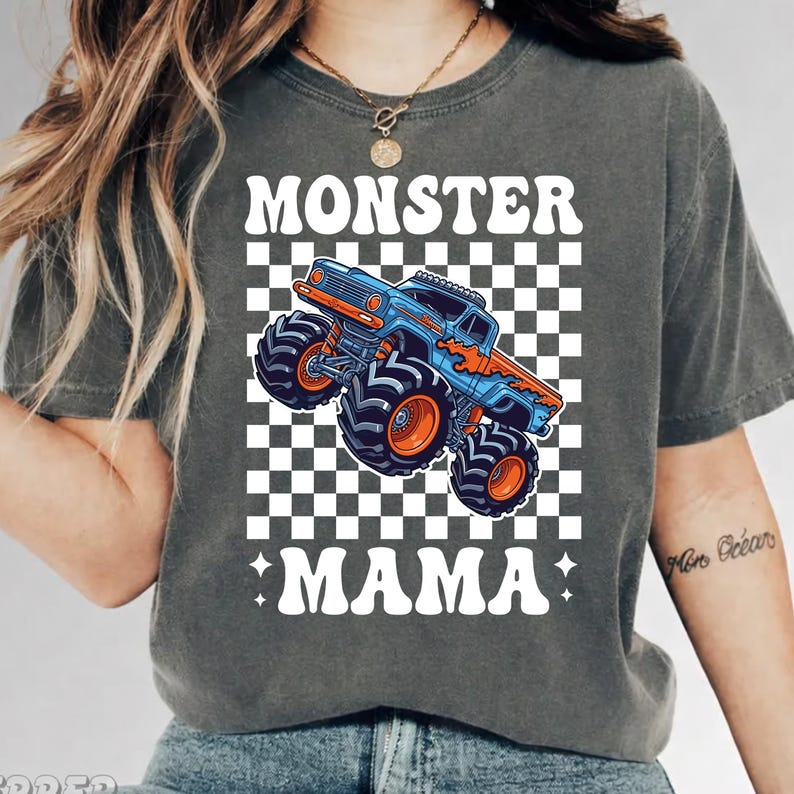 Op de afbeelding: Donkergrijs T-shirt met een blauwe en oranje monstertruck op een zwart-wit geblokte achtergrond. De tekst "MONSTER MAMA" is in het wit gedrukt, met een ster aan weerszijden van het woord "MAMA".
