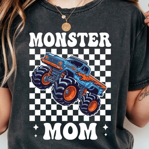 Puede incluir: Camiseta negra con un dise&ntilde;o de cami&oacute;n monstruo azul y naranja sobre un fondo a cuadros. Las palabras "MONSTER MOM" est&aacute;n impresas en blanco con una fuente estilizada.