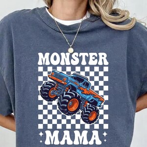 Op de afbeelding: Een marineblauw sweatshirt met een afbeelding van een blauwe en oranje monstertruck op een geruite achtergrond. De woorden "MONSTER MAMA" staan in het wit boven en onder de truck.