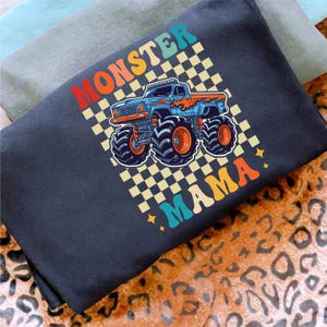 May include: A navy blue t-shirt with a colorful graphic of a monster truck. The text "MONSTER MAMA" is displayed in a retro font. The truck is blue with orange accents and large tires, set against a checkered background.