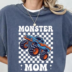 Puede incluir: Una sudadera azul marino con un gr&aacute;fico de un cami&oacute;n monstruo azul y naranja sobre un fondo a cuadros. Las palabras "MONSTER MOM" est&aacute;n impresas en letras blancas en la parte superior e inferior del cami&oacute;n.