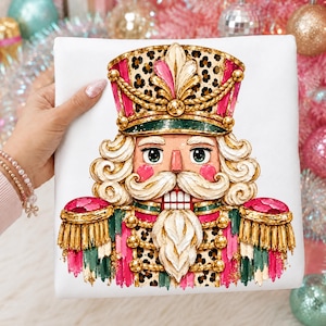 May include: A white sweatshirt featuring a graphic print of a nutcracker soldier. The nutcracker wears a leopard print hat with gold trim, a green and pink uniform with gold tassels, and has a white beard and mustache.