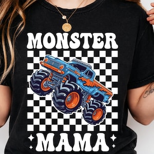 Op de afbeelding: Zwart T-shirt met een afbeelding van een blauwe en oranje monstertruck. De tekst "MONSTER MAMA" staat in het wit boven en onder de truck, tegen een zwart-witte geblokte achtergrond. Het shirt heeft een ronde hals.