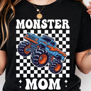 Puede incluir: Camiseta negra con las palabras "MONSTER MOM" en blanco, con un gr&aacute;fico de cami&oacute;n monstruo azul y naranja sobre un fondo a cuadros. El dise&ntilde;o est&aacute; centrado en la camiseta.