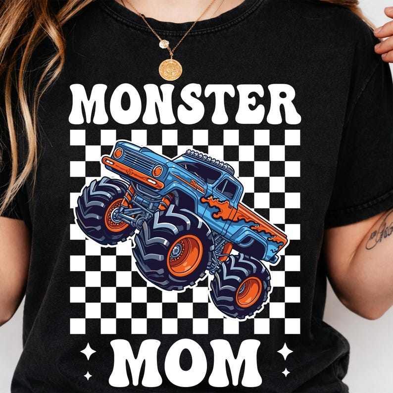Puede incluir: Camiseta negra con las palabras "MONSTER MOM" en blanco, con un gr&aacute;fico de cami&oacute;n monstruo azul y naranja sobre un fondo a cuadros. El dise&ntilde;o est&aacute; centrado en la camiseta.