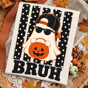 May include: A cream-coloured fabric featuring a graphic of a white ghost wearing a black baseball cap and sunglasses, holding an orange jack-o'-lantern. The design includes black vertical stripes with white polka dots and the word BRUH in black.