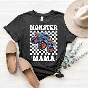 Op de afbeelding: Donkergrijs t-shirt met een blauw-oranje monstertruck-afbeelding op een zwart-witte geblokte achtergrond. De tekst "MONSTER MAMA" staat boven en onder het truckontwerp. Er zijn ook beige ballerina's te zien.