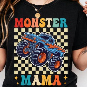 May include: Black t-shirt with a colorful graphic of a monster truck. The text "MONSTER MAMA" is displayed in a retro font above and below the truck. The truck is blue and orange, set against a checkered background.
