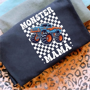Op de afbeelding: Zwart T-shirt met een blauwe en oranje monstertruck op een geruite achtergrond. De woorden "MONSTER MAMA" zijn in het wit gedrukt. Het shirt is opgevouwen, met andere shirts zichtbaar op de achtergrond.
