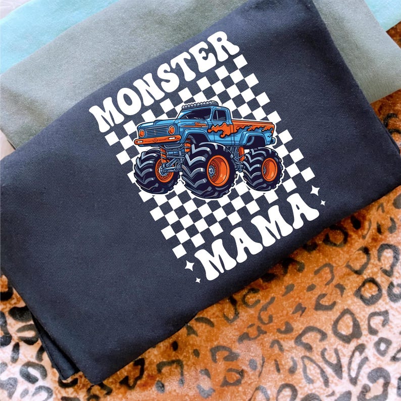 Op de afbeelding: Zwart T-shirt met een blauwe en oranje monstertruck op een geruite achtergrond. De woorden "MONSTER MAMA" zijn in het wit gedrukt. Het shirt is opgevouwen, met andere shirts zichtbaar op de achtergrond.