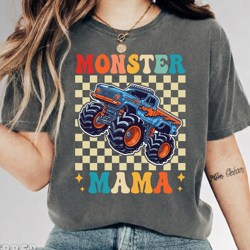 May include: Dark gray t-shirt featuring a colorful graphic of a monster truck with the words "MONSTER MAMA" in a retro font. The design includes a checkered background and star accents, creating a vintage, playful aesthetic.