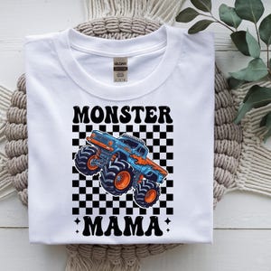 May include: White t-shirt with a graphic of a blue and orange monster truck on a black and white checkered background. The words "MONSTER MAMA" are printed above and below the truck in black.