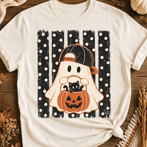 May include: A cream-coloured t-shirt featuring a graphic of a ghost wearing a black baseball cap. The ghost holds an orange jack-o'-lantern bucket containing a small black cat. The design is set against a background of black vertical stripes with white polka dots.
