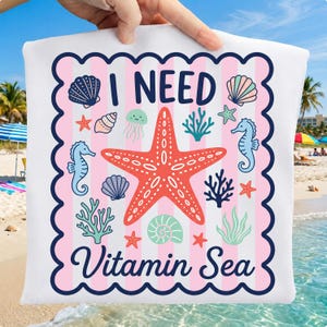 May include: White pillow with a pink and white striped background, featuring a red starfish and various sea creatures. The text "I NEED Vitamin Sea" is displayed in navy blue script, perfect for beach decor.