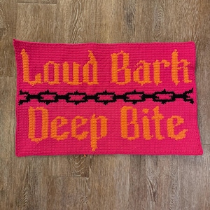 May include: A rectangular, hot pink crochet piece with the words "Loud Bark" and "Deep Bite" in orange, gothic-style lettering. A black chain design separates the phrases. The item is displayed on a wooden surface.