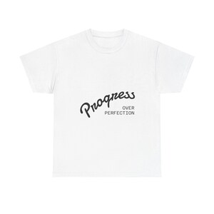 Può includere: T-shirt bianca con la scritta "Progress Over Perfection" in nero, in corsivo e con caratteri senza grazie. La maglietta è di stile girocollo, adatta per un uso casual.