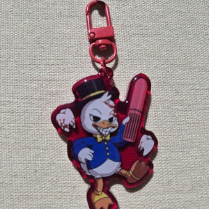 May include: A red acrylic keyring featuring a cartoon duck character wearing a black top hat, a blue jacket, and a yellow bowtie. The character holds a pink lipstick and has a tooth floating nearby, with red accents throughout the design.