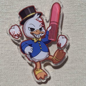 May include: A clear acrylic charm featuring a cartoon duck character wearing a black top hat, a blue jacket, and a yellow bow tie. The duck holds a pink cylindrical object and is surrounded by two white teeth with red blood-like splatters.