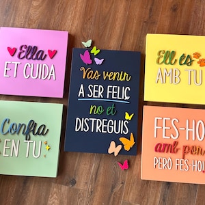May include: A collection of six colorful square wooden plaques with inspirational phrases in Catalan. The plaques are in various colors, including pink, green, yellow, orange, and navy blue. Some plaques have butterfly decorations. The text includes phrases like "Ella ET CUIDA" and "Confia EN TU".