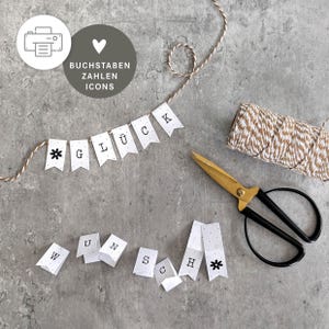 May include: A DIY banner kit featuring white paper pennants with black letters spelling GLÜCK WUNSCH and flower icons. The set includes a spool of brown and white twisted twine and a pair of gold and black scissors for assembly and customization.