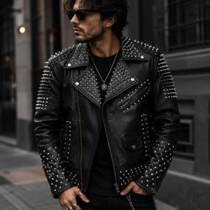 May include: Black leather jacket adorned with silver metal studs on the shoulders, sleeves, and lapels. The jacket features a classic biker design with a zip closure and pockets. A silver chain hangs from the pocket. The jacket is worn with a black shirt and sunglasses.
