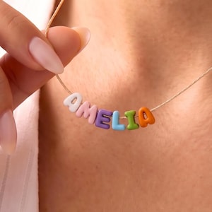 May include: A delicate necklace featuring a thin gold-toned chain with colorful, rounded letter beads spelling out the name AMELIA. The beads are white, pink, purple, blue, green, and orange, creating a vibrant, playful, and personalized accessory.